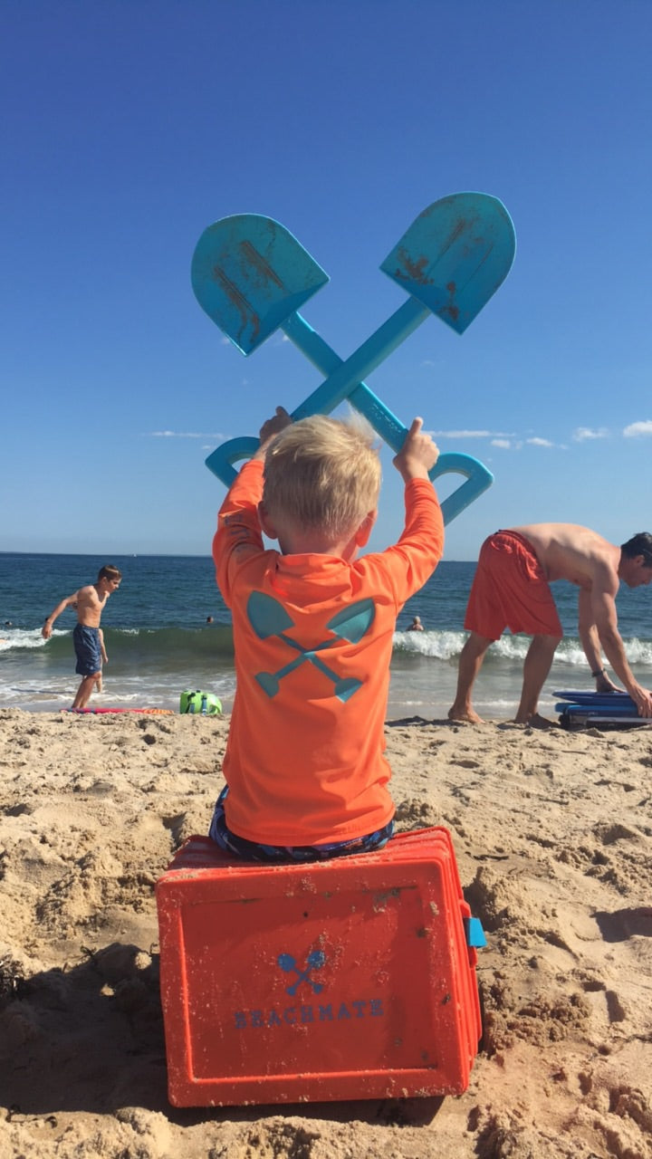 Child in orange long‑sleeve rash guard with sun hat holding a shovel at the beach