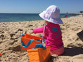 Load image into Gallery viewer, A baby wearing a white sun hat with a polka dot pattern, sitting on a beach and playing with beach toys.
