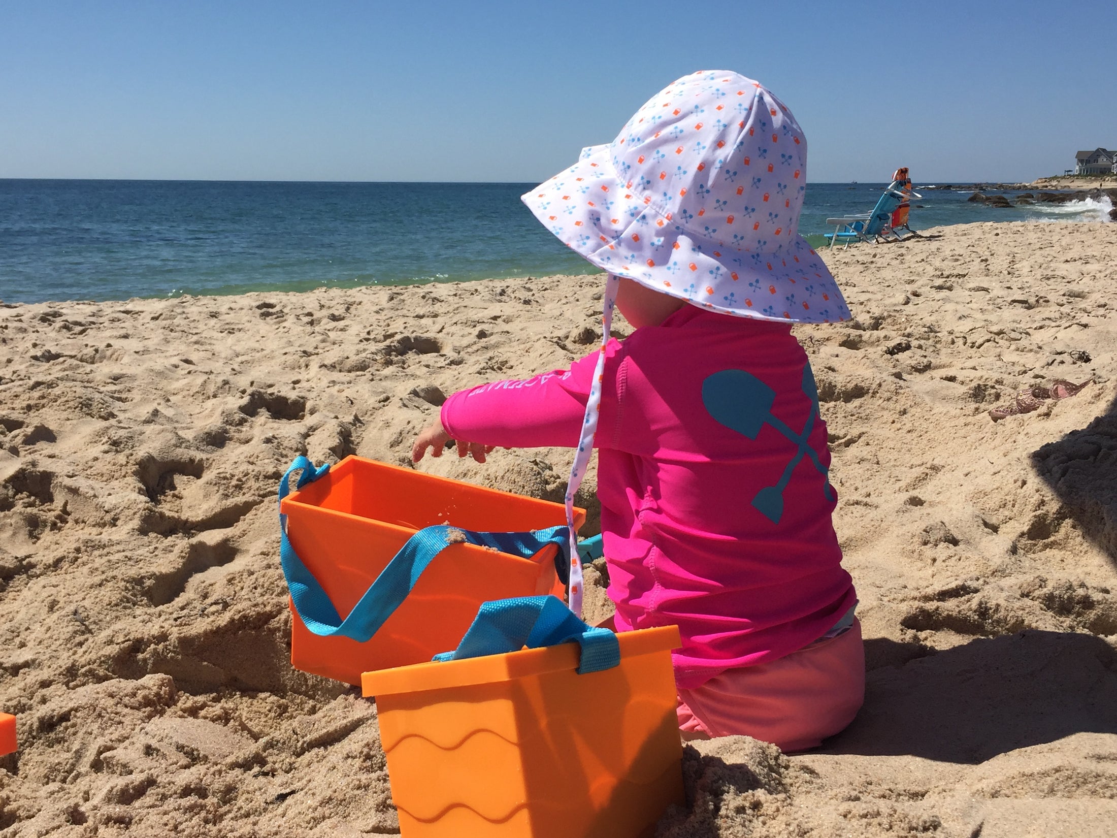 A baby wearing a white sun hat with a polka dot pattern, sitting on a beach and playing with beach toys.
